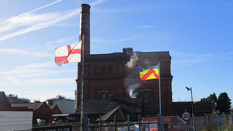 Façade of Victorian building. 2 flags are flying at the entrance. Steam rises from a low building.