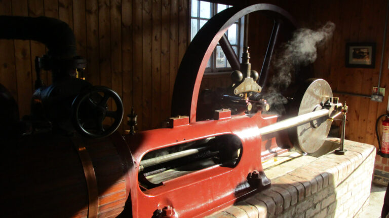 Cylinder of steam engine in foreground, with wooden slatted cladding. Crank and flywheel in the background.