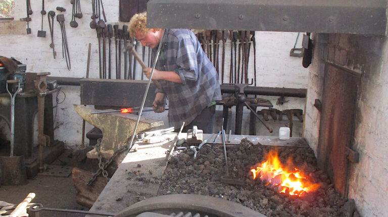 Blacksmith hammers red-hot metal on an anvil as he re-shapes the end of a pair of tongues